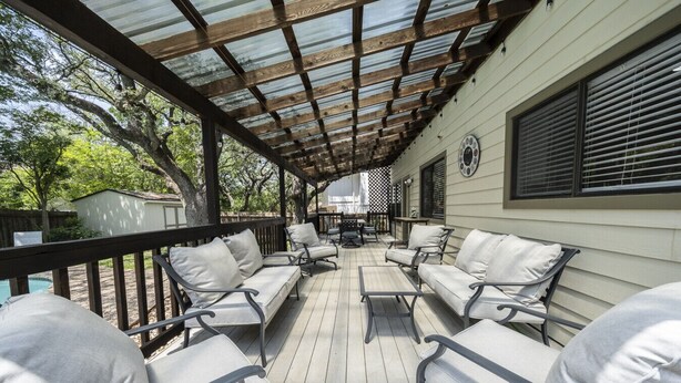 A covered outdoor seating area with cushioned chairs and a table, a wooden deck, and a building with windows and a door.