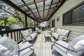 Un espace extérieur abrité avec des chaises rembourrées et une table, une terrasse en bois et un bâtiment doté de fenêtres et d’une porte.
