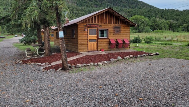A rustic cabin named Fort Wilderness with a wooden exterior, a porch with red chairs, and a gravel driveway. - Historic Ft Wilderness Cabin (South Fork)