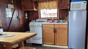 A compact kitchen with wooden cabinets, a stove, a refrigerator, and a window with a view. - Historic Ft Wilderness Cabin (South Fork)