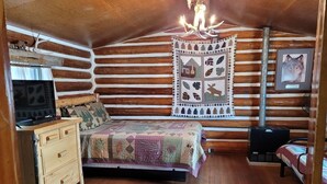 A log cabin bedroom with a bed, a dresser, a television, a quilt, a rug, and a fireplace. - Historic Ft Wilderness Cabin (South Fork)