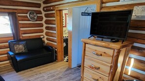 L’intérieur d’un chalet accueillant, avec un plancher en bois, un canapé noir, une commode en bois et un téléviseur posé sur un meuble en bois.