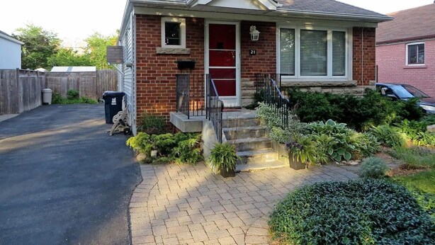 A single-story house with a brick facade, a front porch with steps, a mailbox, and a small garden area with various plants.