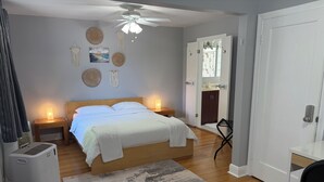 Un dormitorio con cama, lámparas de noche, ventilador de techo, una silla, escritorio y una puerta que da a un baño.