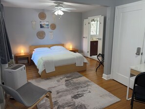 A bedroom with a bed, bedside lamps, a ceiling fan, a chair, a desk, and a door leading to a bathroom.
