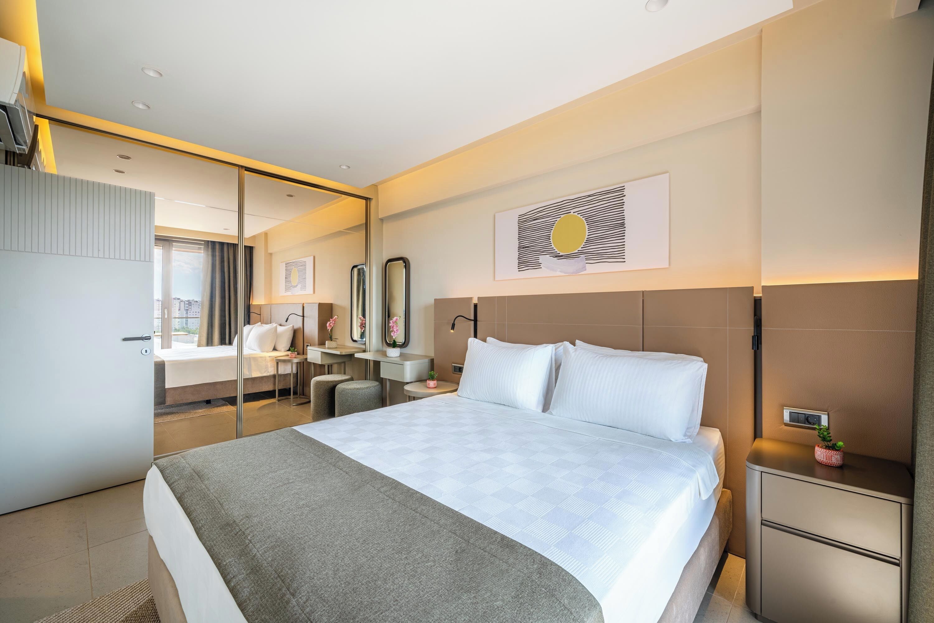 A modern hotel room with a large bed, a desk, a chair, a mirror, and a small plant on the nightstand.