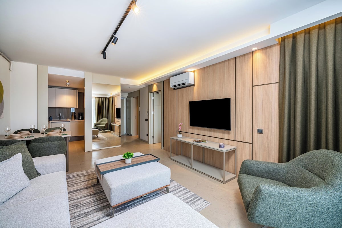 A modern living room with a flat-screen TV, green upholstered sofas, a coffee table, and a dining area with a kitchen in the background.
