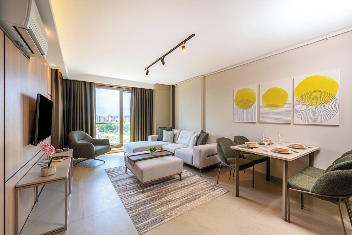 A modern hotel room with a sofa, a dining table set for two, a flat-screen TV, and a large window with curtains.