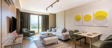A modern hotel room with a sofa, a dining table set for two, a flat-screen TV, and a large window with curtains.