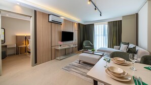 A modern hotel room with a flat-screen TV, a sofa, a dining table set with plates and glasses, a small side table, and a large window with curtains. - Casamax Suites (Konyaalti)