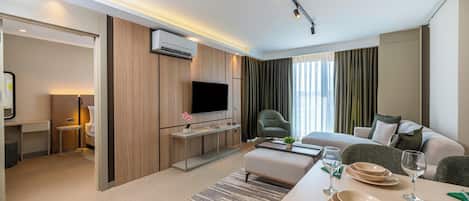 A modern hotel room with a flat-screen TV, a sofa, a dining table set with plates and glasses, a small side table, and a large window with curtains.