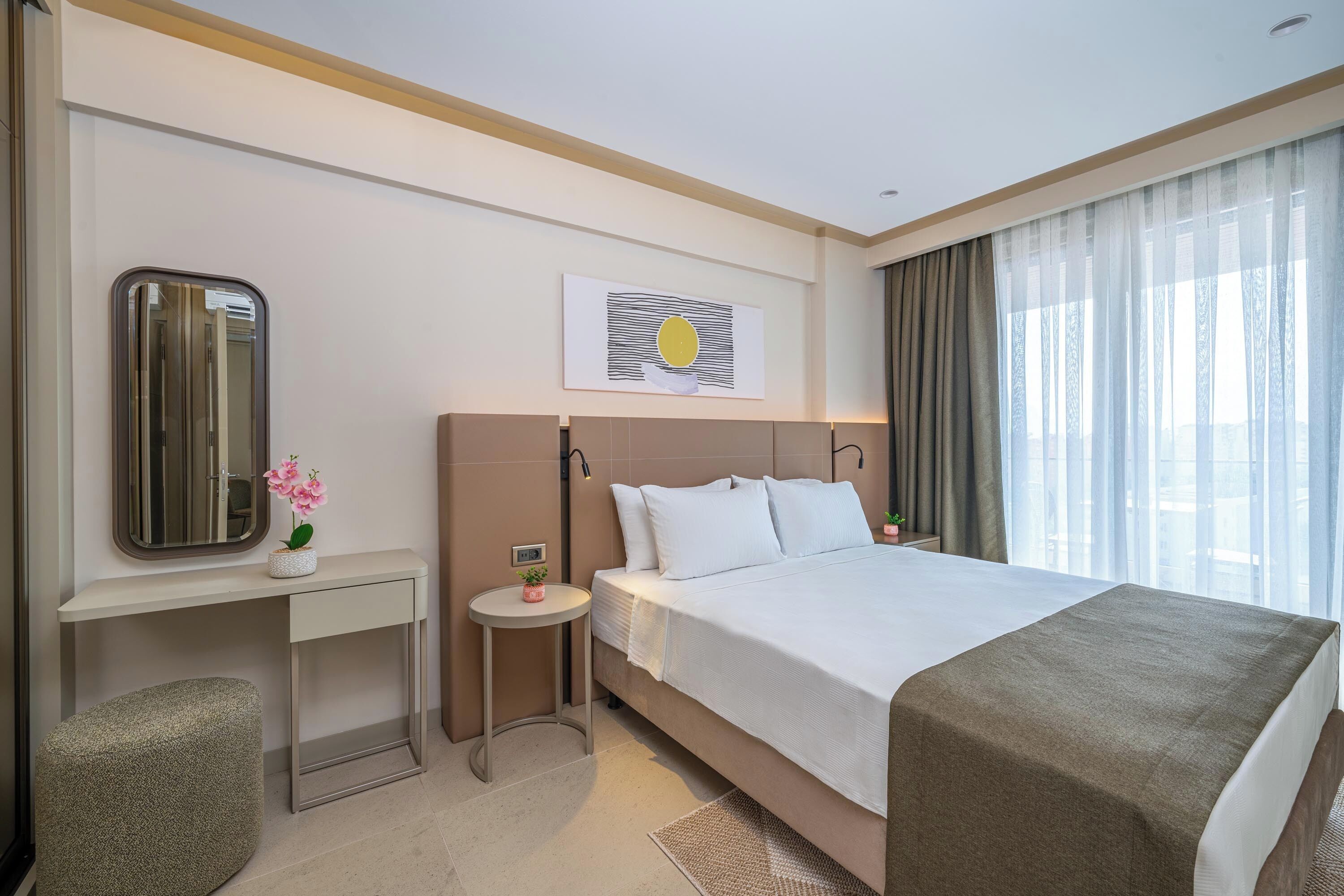 A hotel room with a large bed, a desk, a small table with a plant, a mirror, and a picture on the wall.