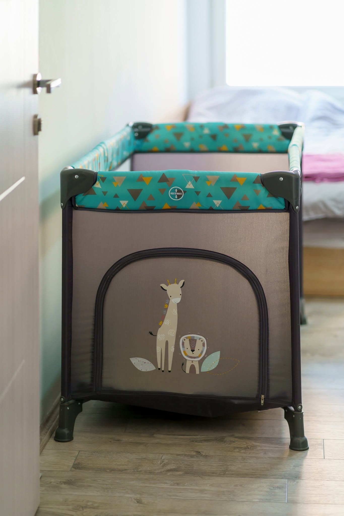 A portable crib with a giraffe design, positioned in a room with a wooden floor and a bed in the background.