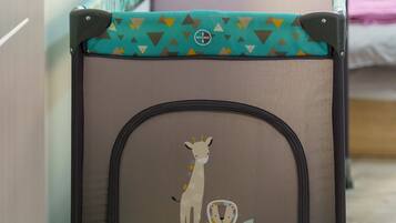 Un berceau portable à motifs de girafe, placé dans une pièce dont le sol est en bois et où l’on aperçoit un lit au fond.