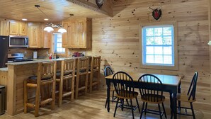 Interior de una cabaña de madera con cocina, comedor y ventana con persianas.