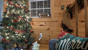 Interior acogedor de una cabaña con un árbol de Navidad decorado, mantas de cuadros y una almohada que dice 'Merry Christmas'.