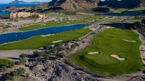 Ein Golfplatz mit einem Hotel im Hintergrund, umgeben von Bergen und einem Gewässer.