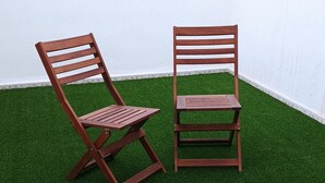 Zwei Holzstühle mit Lamellenlehnen und -sitzen auf künstlichem Grün vor einer weißen Wand.