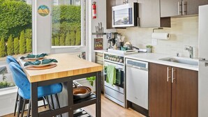 Moderni keittiö, jossa on puinen pöytä, siniset tuolit, ruostumattomasta teräksestä valmistetut kodinkoneet ja näkymä vehreään maisemaan.