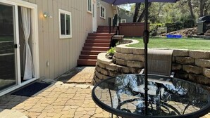 A patio area with a glass table and a purple umbrella, a stone retaining wall, and a wooden deck with steps leading to a door. - Luxury 4 Bedroom + Hot Tub. (El Dorado Hills)