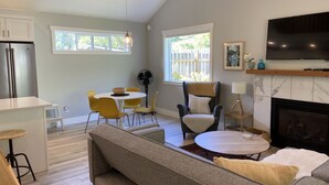 A modern living room with a sofa, a dining table with chairs, a kitchen area with a refrigerator, and a fireplace. - Roberts Creek Cottage. Close to village, beach, hiking and biking trails. (Roberts Creek)