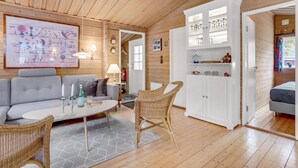 A cozy room with a grey sofa, wicker chairs, a white cabinet with glassware, and a wooden ceiling.
