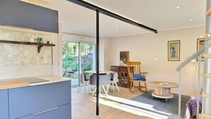 A modern kitchen with a sliding door leading to a dining area. There is a wooden dining table with chairs, a small round table, and a rocking chair. The room has a staircase with a metal railing. - L'optimist - Pluneret (PLUNERET)