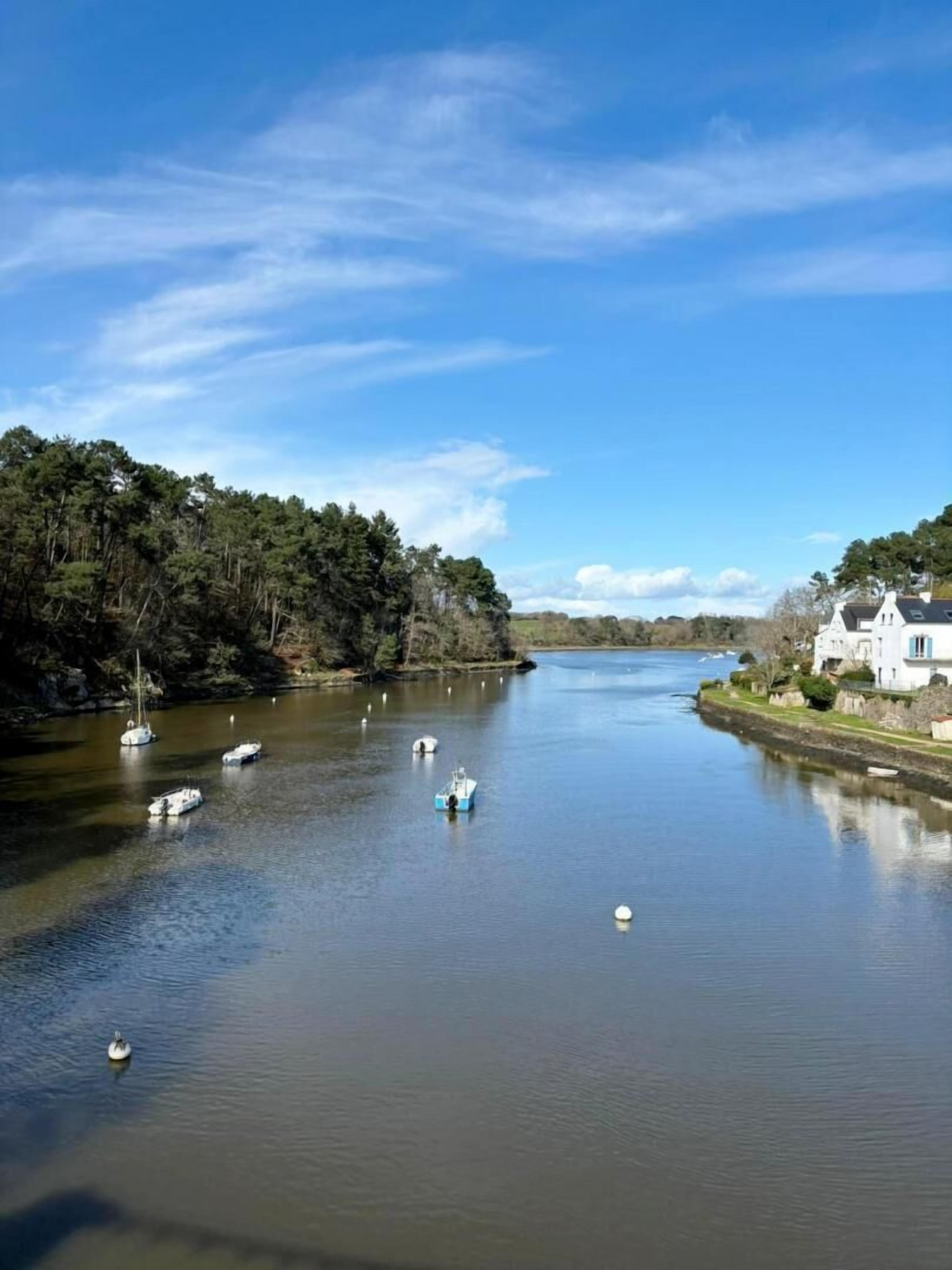 Une rivière paisible où naviguent des bateaux, bordée d’arbres denses et dominée par un bâtiment blanc au toit en pente.