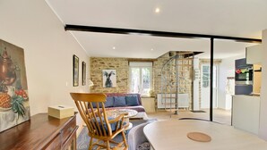 A modern living room with a wooden rocking chair, a sofa, and a dining table. There is a staircase leading to an upper level, and a kitchen area is visible in the background. - L'optimist - Pluneret (PLUNERET)