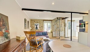 Uma sala de estar moderna com uma cadeira de balanço de madeira, um sofá e uma mesa de jantar. Uma escada leva a um andar superior, e uma área de cozinha é visível ao fundo.