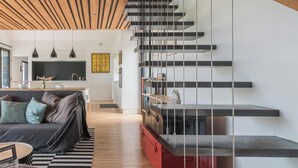 Un intérieur moderne avec un plafond en bois, un escalier doté de rampes en métal, un coin cuisine et un espace de vie comprenant un canapé et des coussins.