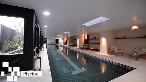 A long indoor swimming pool with a tiled floor, surrounded by lounge areas with chairs and tables, and a large window on one side. - Kazanoah - Campo Belo (São Paulo)