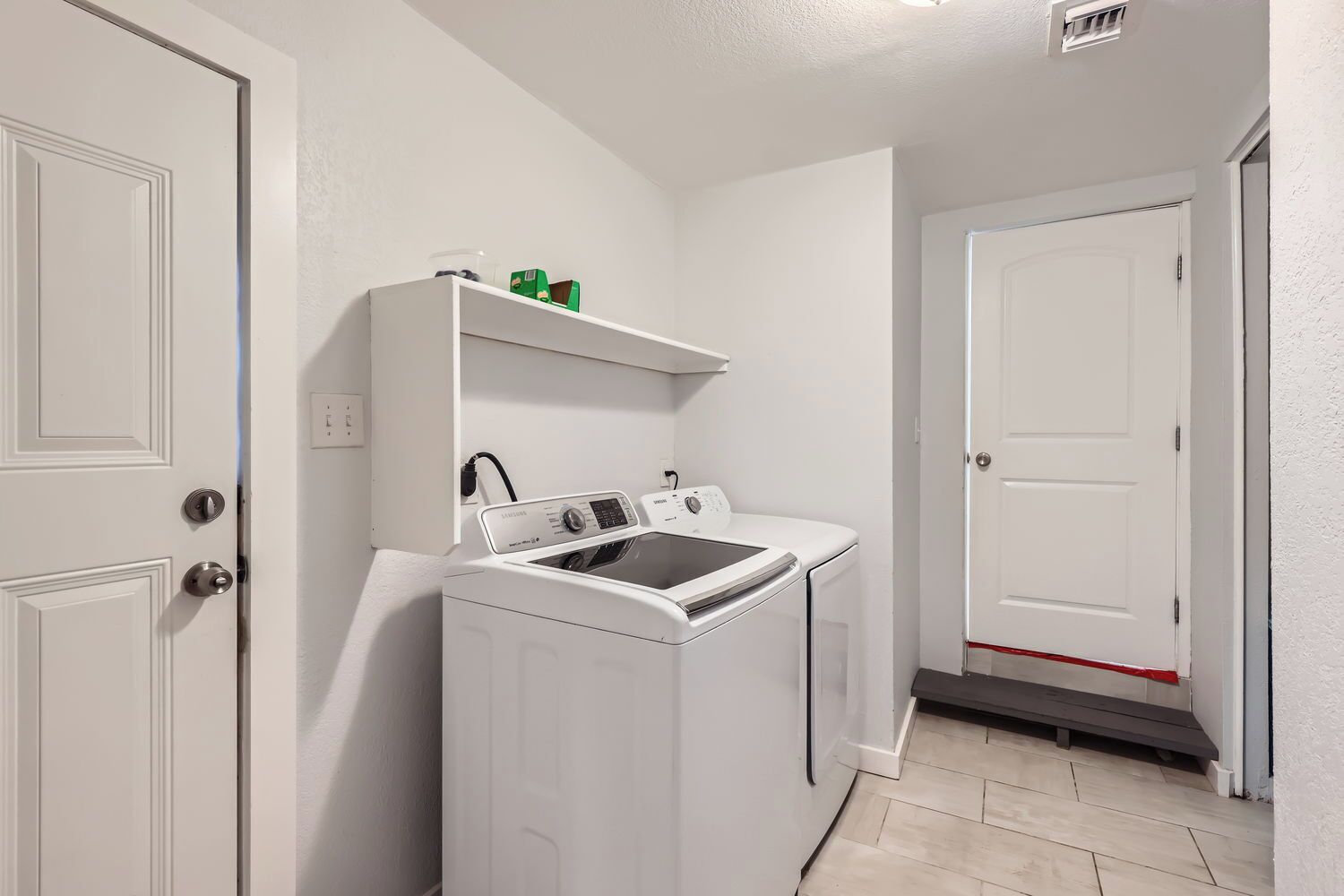 A laundry room with a front-loading washer and dryer, a shelf above the dryer, and a door leading to another room.