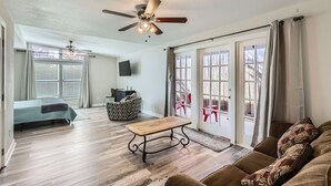 A spacious living room with a wooden floor, a ceiling fan, a sofa, a coffee table, and a sliding glass door leading to an outdoor area with red chairs.