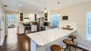 A modern kitchen with white cabinets, stainless steel appliances, a marble island with bar stools, pendant lighting, and a chalkboard with a list.