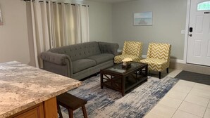 Una sala de estar con un sofá gris, dos sillones con cojines de estampado geométrico, una mesa de centro de vidrio y una zona de cocina con encimera de mármol.