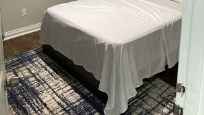 Un dormitorio con cama, alfombra estampada, pared de color claro y un cuadro con plantas.
