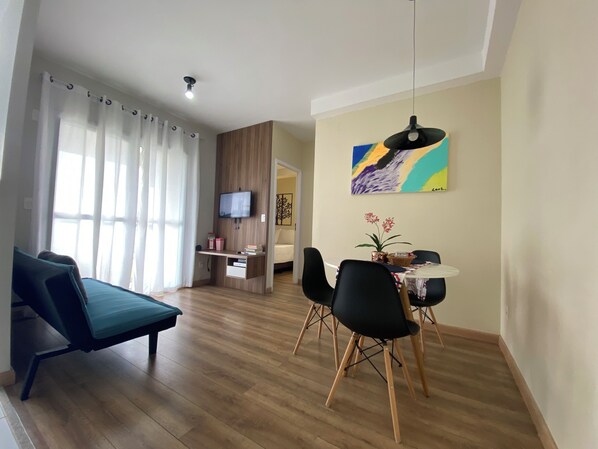 A modern living room with a sofa, a dining table with chairs, a TV mounted on the wall, and a painting above the table.