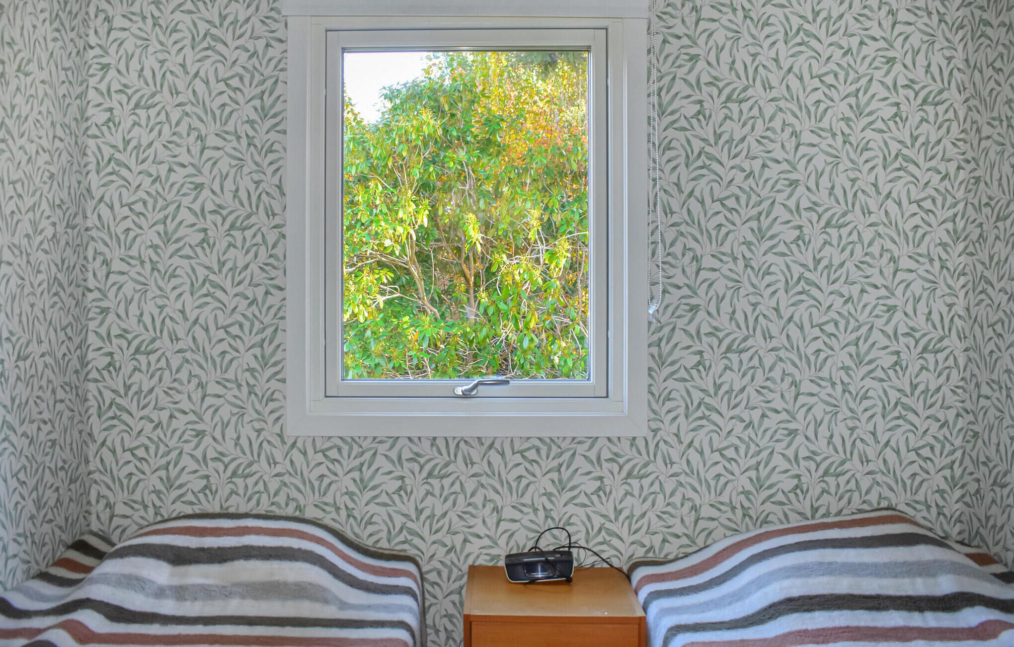Ein Schlafzimmer mit Fenster, strukturiertem Wandpapier, zwei Betten und einem kleinen Nachttisch mit Radio.