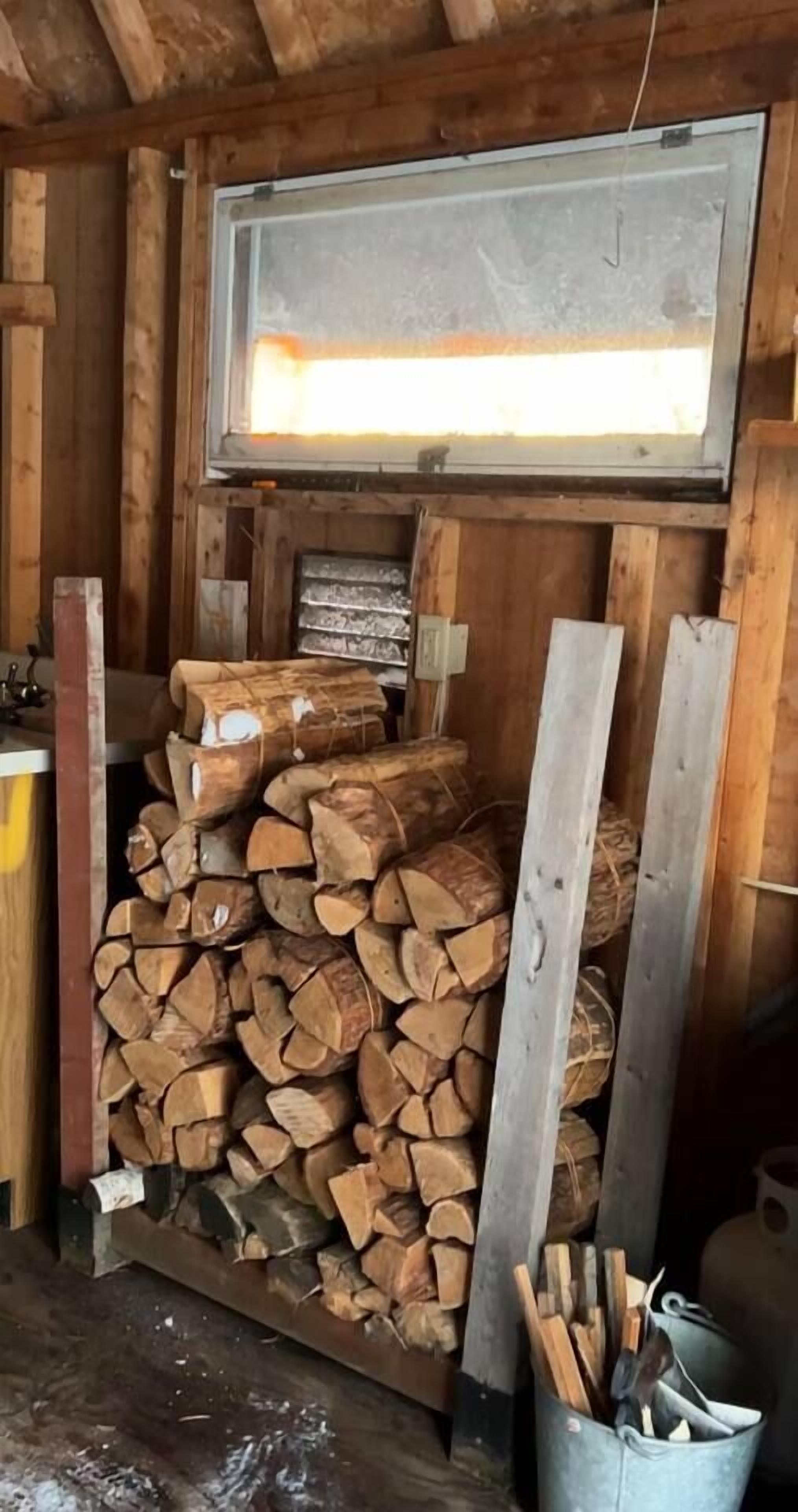 A rustic wooden cabin interior with a stack of firewood, a metal bucket with tools, and a window above the woodpile.