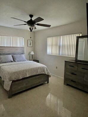 Une chambre à coucher comprenant un lit, une table de chevet, une commode, un ventilateur de plafond et une fenêtre avec des stores.