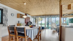 Ein Essbereich mit Holzbalkendecke, einem runden Tisch mit Stühlen, einer Küchenzeile mit Grill und großen Fenstern mit Blick ins Freie.