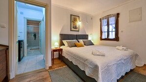 A bedroom with a bed, a nightstand, a window with curtains, a painting on the wall, and a bathroom visible through an open door.