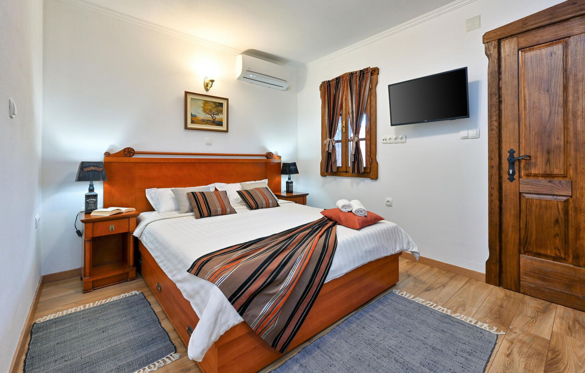 A neatly arranged bedroom with a wooden bed, a flat-screen TV, a wooden wardrobe, and a bedside table with a lamp.