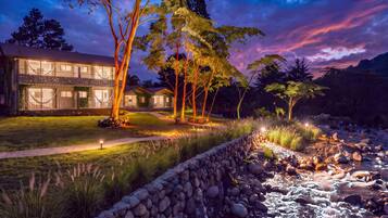 A modern house with a stone facade, a well-maintained lawn, and a stone retaining wall beside a flowing stream.