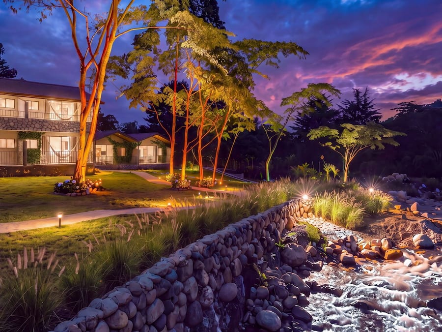 A modern house with a stone facade, a well-maintained lawn, and a stone retaining wall beside a flowing stream.