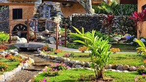 Un jardín con un puente de piedra, una cascada pequeña y diversas plantas y flores.