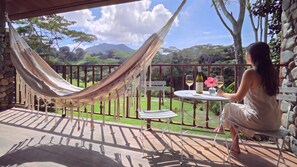 A hammock on a balcony with a table and chairs, overlooking a lush garden and distant mountains.