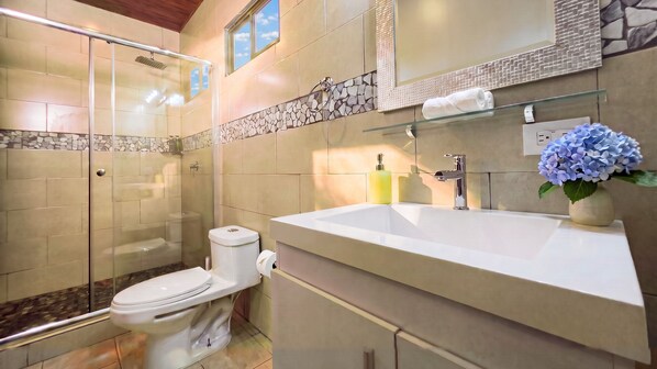 A modern bathroom with a glass shower enclosure, a white toilet, a large sink with a mirror above, and a vase with blue flowers.