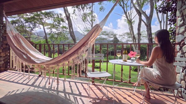 A hammock chair and a table with a bottle and glasses on a balcony with a view of trees and mountains.